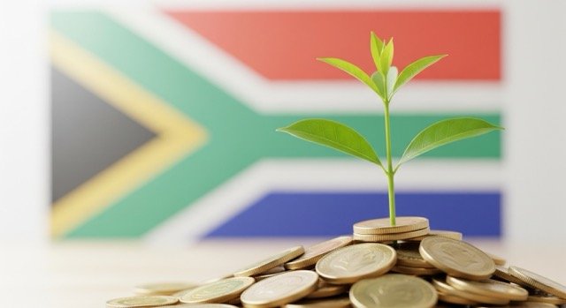 A landscape image depicting a small green plant sprouting from a pile of shiny South African coins, symbolizing investment and savings growth. The South African flag is visible in the soft-focus background.